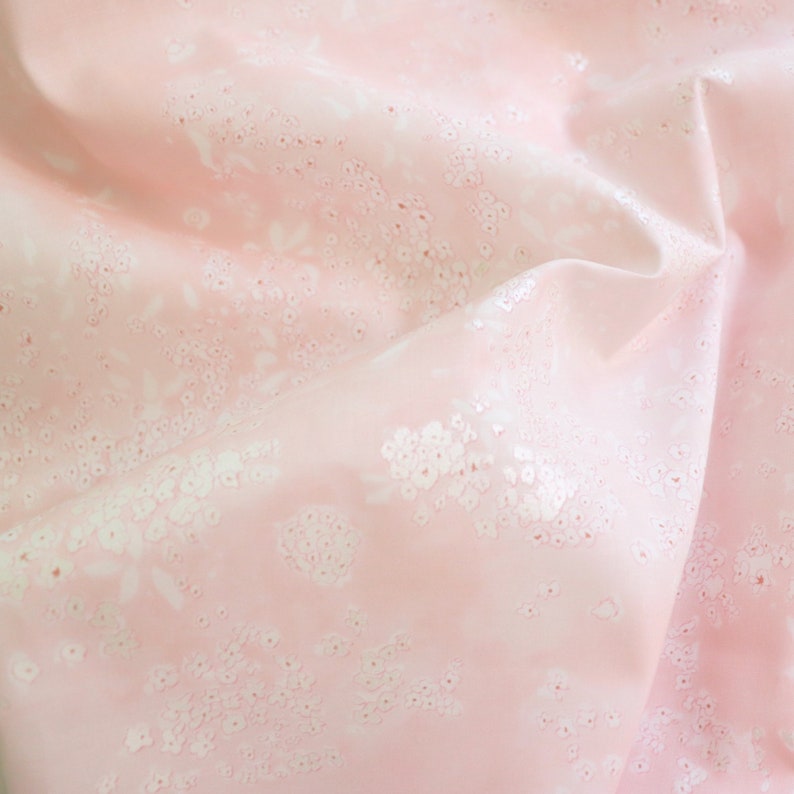 Pode incluir: Close-up de um tecido rosa com um delicado padr&atilde;o floral. O tecido tem uma apar&ecirc;ncia macia e ligeiramente brilhante, com desenhos florais e de folhas brancas espalhados pela superf&iacute;cie. O tecido est&aacute; dobrado, criando sombras e profundidade.