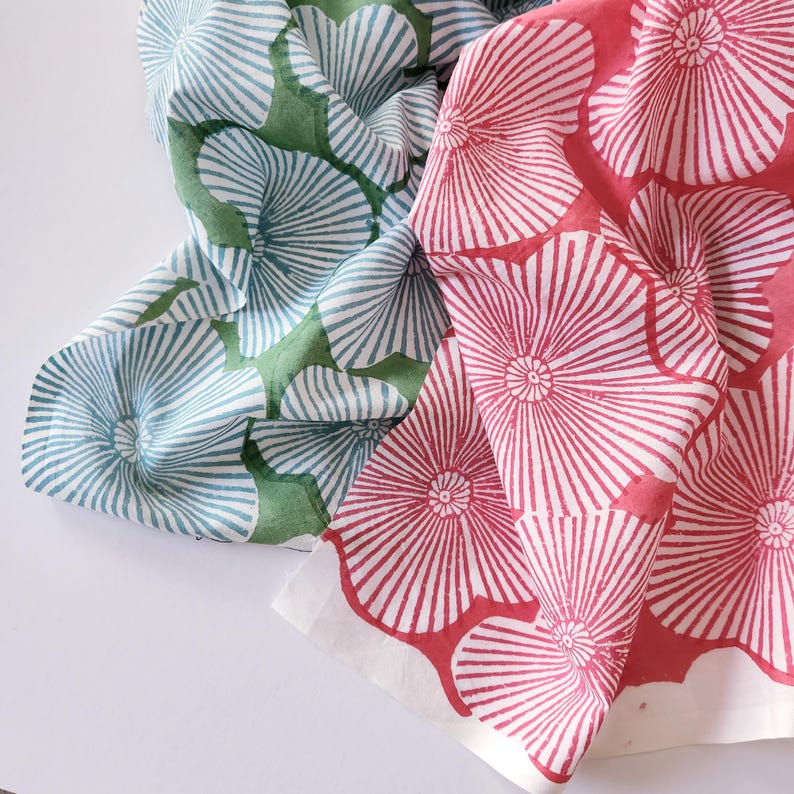 May include: Two pieces of fabric featuring a repeating block-printed floral pattern. One piece displays light blue flowers on a green background, while the other features vibrant pink flowers on a white background, both with radiating petal lines.
