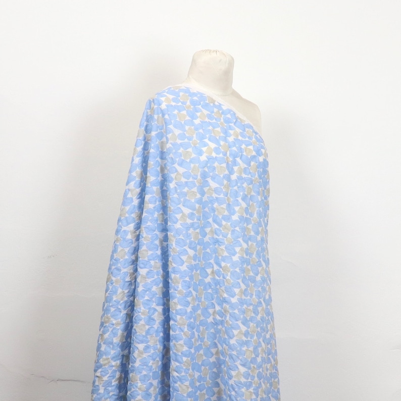 May include: A bolt of fabric draped over a mannequin. The fabric features a pattern of blue and gray shapes on a white background. The fabric is a light, airy material, suitable for clothing or home decor.