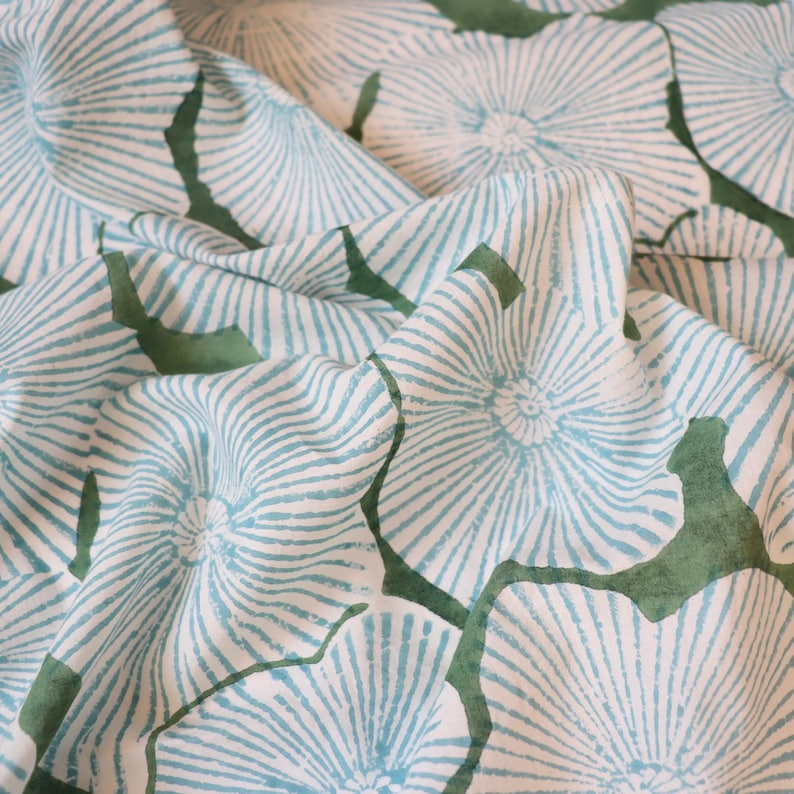 May include: A white fabric featuring a repeating pattern of light blue, radiating, fan-like floral shapes with dark green, organic, leaf-like silhouettes interspersed between the flowers, creating a textured, folded appearance across the surface.