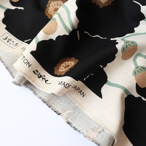 May include: Close-up of a fabric with a floral pattern. The design features large black flowers, brown accents, and green stems on a cream background. The fabric has text that reads "COTTON" and "MADE IN JAPAN".