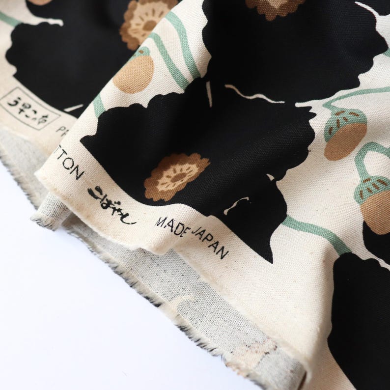 May include: Close-up of a fabric with a floral pattern. The design features large black flowers, brown accents, and green stems on a cream background. The fabric has text that reads "COTTON" and "MADE IN JAPAN".