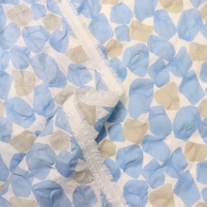 May include: A folded piece of fabric with a repeating pattern of blue and tan shapes on a white background. The fabric has a delicate, slightly wrinkled texture and a finished edge with small holes.