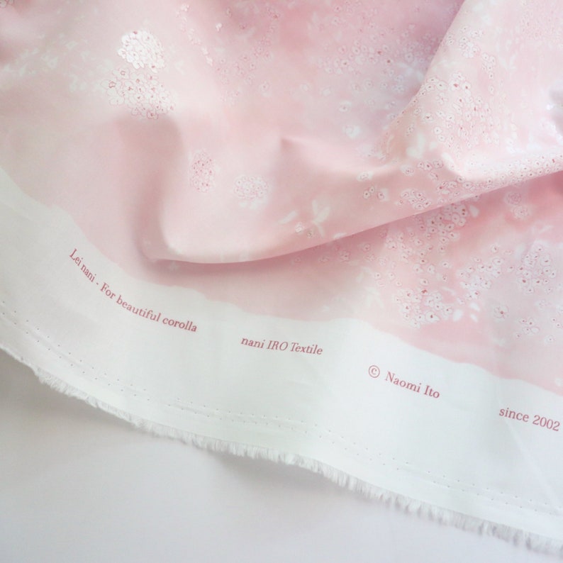 Pode incluir: Tecido rosa claro com um padr&atilde;o floral e o texto "Lei nani - For beautiful corolla" e "nani IRO Textile". O tecido tem um tom rosa suave com detalhes brancos e uma borda branca. O texto "&copy; Naomi Ito since 2002" tamb&eacute;m &eacute; vis&iacute;vel.