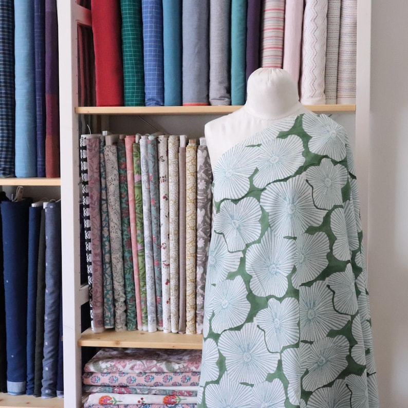 May include: A white dress form draped with green fabric featuring a large white floral pattern. The background consists of a wooden shelving unit filled with numerous rolls of fabric in various colors, textures, and patterns arranged vertically.