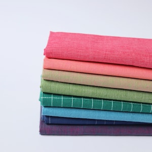 May include: A stack of colorful fabric swatches in shades of pink, green, blue, and purple. The fabrics have a textured appearance, with some featuring a grid pattern. The swatches are neatly stacked, showcasing a variety of colors and textures.