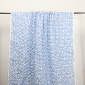 May include: A light blue and white quilt with a pattern of small, irregular shapes in light blue and gray. The quilt is hanging on a metal rod against a white background. The edges of the quilt are white.