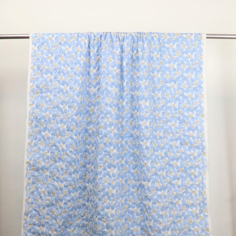 May include: A light blue and white quilt with a pattern of small, irregular shapes in light blue and gray. The quilt is hanging on a metal rod against a white background. The edges of the quilt are white.