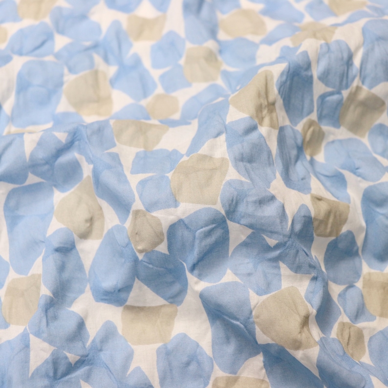 May include: Close-up of a white fabric with a pattern of blue and tan irregular shapes. The fabric is slightly wrinkled, creating shadows and depth. The shapes resemble abstract stones or pebbles.
