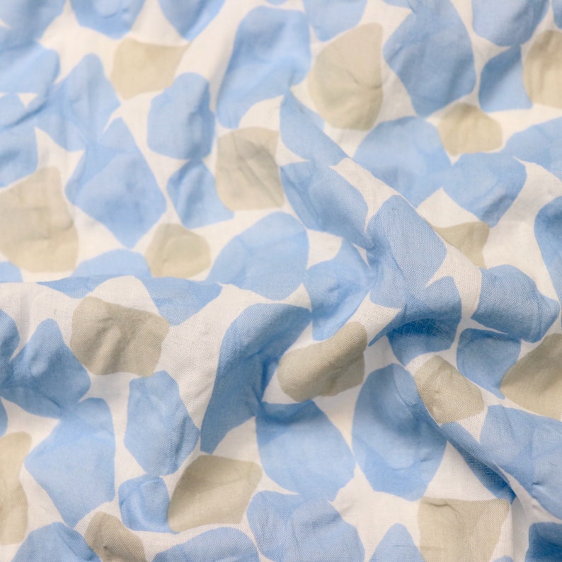 May include: Close-up of a white fabric with a pattern of irregular shapes in light blue and gray. The fabric is slightly wrinkled, creating shadows and texture. The shapes are scattered across the surface.