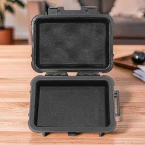 May include: An open, dark gray, rectangular, waterproof case with a black foam insert. The case is on a wooden surface. The case has a hinged lid and a latch closure. The interior is lined with a black foam.