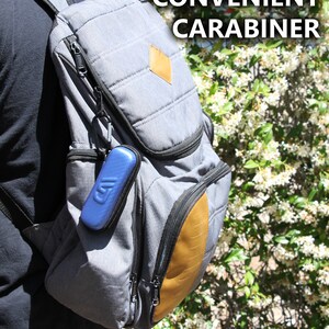 K&ouml;nnte beinhalten: Ein grauer Rucksack mit einem braunen Lederflicken und einem blauen Karabiner, der am Rei&szlig;verschluss befestigt ist. Der Text "CONVENIENT CARABINER" ist in wei&szlig;en Buchstaben auf schwarzem Hintergrund geschrieben.