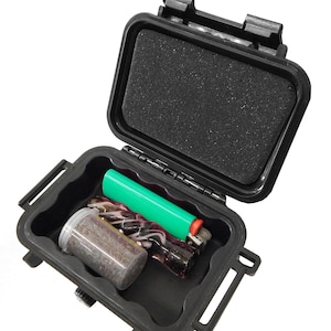 May include: A black rectangular hard-shell storage case with a hinged lid and a foam-lined interior. Inside the open case sits a small clear plastic container filled with dark particles, a green lighter, and a dark patterned glass object.