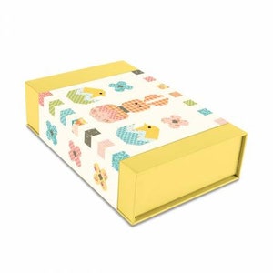 May include: A yellow gift box with a white lid featuring a colourful pattern of bunnies, chicks, and flowers. The box is decorated with a patchwork design and has a spring theme.