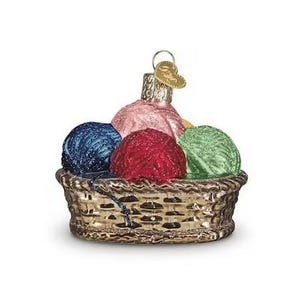May include: A Christmas tree ornament depicting a basket of yarn balls. The basket is gold-coloured with a textured pattern. The yarn balls are blue, red, green, and pink. A gold heart-shaped tag hangs from the top of the ornament.