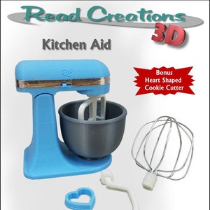 Puede incluir: Un robot de cocina KitchenAid impreso en 3D azul y gris con un accesorio de batidor blanco, un gancho de masa blanco y un cortador de galletas en forma de corazón. La batidora está diseñada para muñecas de 35 a 45 cm. La imagen incluye el texto "Read Creations 3D" y "Bonus Heart Shaped Cookie Cutter".