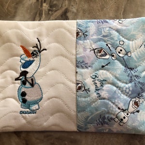 K&ouml;nnte beinhalten: Ein rechteckiger, gesteppter Artikel mit geteiltem Design. Eine Seite ist wei&szlig; mit einem gestickten Olaf-Charakter und dem Wort "Disney". Die andere Seite hat ein blau-wei&szlig;es Muster mit Olaf und den Worten "Ice Breaker!"