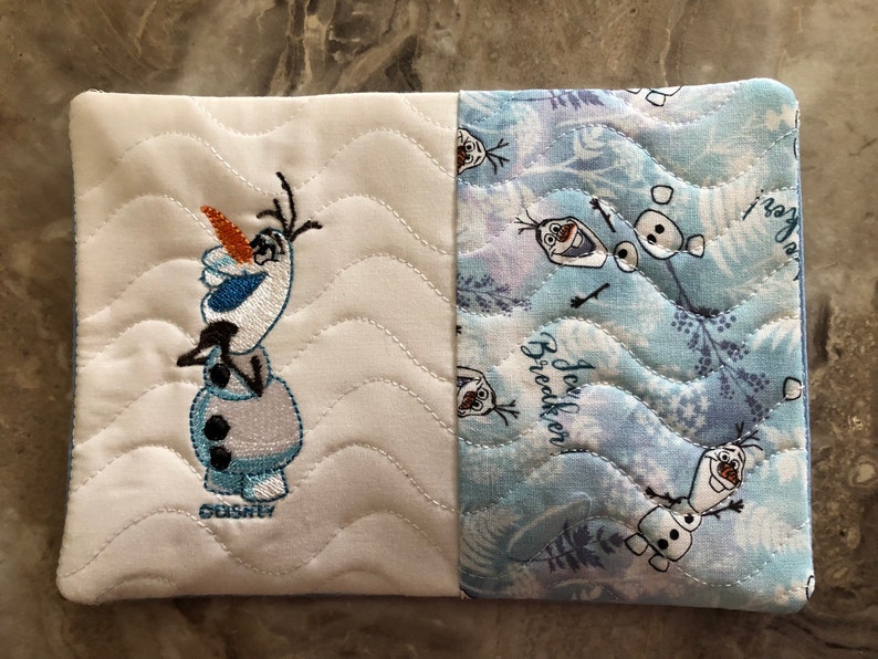 K&ouml;nnte beinhalten: Ein rechteckiger, gesteppter Artikel mit geteiltem Design. Eine Seite ist wei&szlig; mit einem gestickten Olaf-Charakter und dem Wort "Disney". Die andere Seite hat ein blau-wei&szlig;es Muster mit Olaf und den Worten "Ice Breaker!"