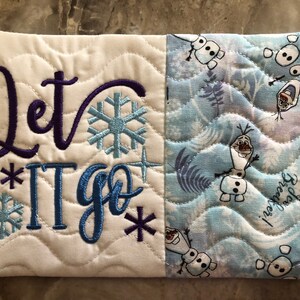 K&ouml;nnte beinhalten: Gestepptes Stoffst&uuml;ck mit dem in Lila und Blau gestickten Schriftzug "Let It Go", zusammen mit Schneeflocken- und Sterndesigns. Die rechte Seite zeigt ein blau-wei&szlig;es Muster mit Olaf dem Schneemann.