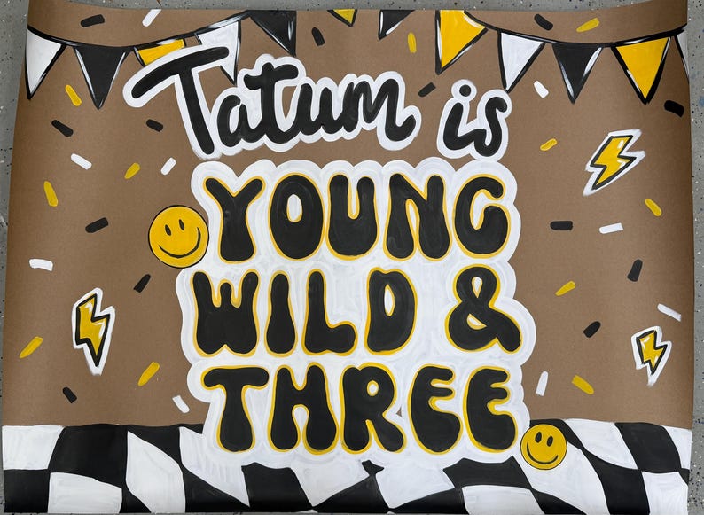 May include: A brown birthday sign with the words "Tatum is YOUNG WILD & THREE" in black and white lettering. The sign has yellow accents, including lightning bolts, confetti, and smiley faces. A black and white checkered pattern borders the bottom.