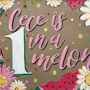 May include: Hand-painted sign with the text "Cece is in a melon" in pink script, with a large white number 1. The sign is decorated with watermelon slices, daisies, and colorful polka dots on a brown background.