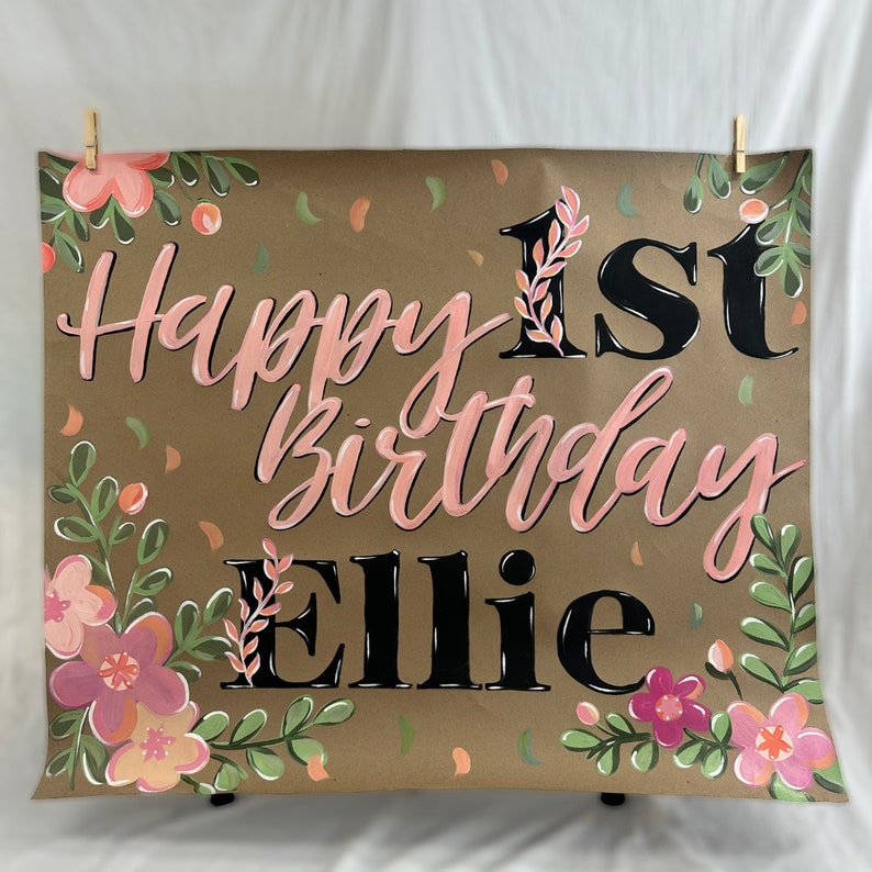 May include: A brown paper sign with the words "Happy 1st Birthday Ellie" in pink and black lettering. The sign is decorated with pink and peach flowers and green leaves. The sign is held up by two clothespins.