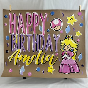 May include: Hand-painted birthday sign on brown paper. The sign says "HAPPY BIRTHDAY Amelia" in pink, purple, and yellow lettering. Features a cartoon princess, mushroom, stars, and confetti. The sign is held up by clothespins.