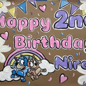 May include: Hand-painted birthday sign on brown paper. The words "Happy 2nd Birthday Nira" are in bubble letters with pink and purple outlines. Features cartoon characters, a rainbow, flowers, hearts, and confetti.