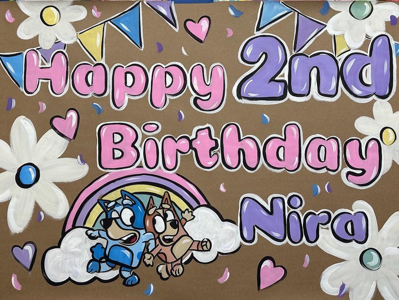 May include: Hand-painted birthday sign on brown paper. The words "Happy 2nd Birthday Nira" are in bubble letters with pink and purple outlines. Features cartoon characters, a rainbow, flowers, hearts, and confetti.