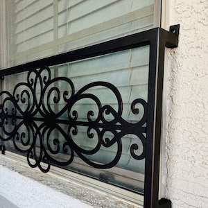 May include: Black metal window security bars with a decorative scroll design. The bars are mounted to the outside of a white stucco wall.