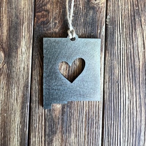 May include: A metal ornament shaped like the state of New Mexico with a heart cut out in the center. The ornament is hanging from a twine string against a wood background.