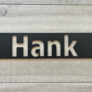 May include: Black metal sign with the name "Hank" cut out. The sign is rectangular with rounded corners and two holes for mounting. The sign is displayed against a light gray wooden background.
