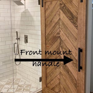 May include: A wooden barn door with a chevron pattern and black hardware. The door is mounted on a black track and has a black handle. The text "Front mount handle" is written on the image.