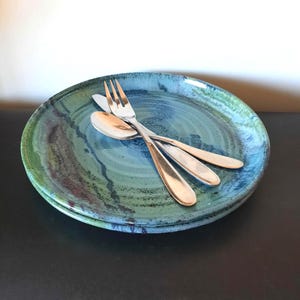 Può includere: Due piatti in ceramica fatti a mano con smalto blu e verde, impilati con una forchetta e due cucchiai. I piatti hanno un aspetto rustico e organico, perfetti per servire i pasti. Le posate sono in acciaio inossidabile.