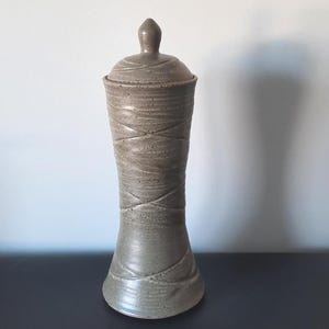 May include: A ceramic jar with a lid, featuring a light brown color and a textured, woven design. The jar has a rounded base and a tall, cylindrical body.