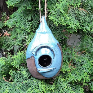 May include: A handmade ceramic birdhouse with a blue glaze and brown accents. The birdhouse has a round opening and is suspended by a natural twine cord. The background is a lush green evergreen tree.