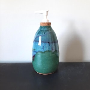 May include: A handmade ceramic soap dispenser with a white pump and cork stopper. The dispenser features a gradient of colours, transitioning from a deep green base to shades of blue and teal at the top. The dispenser is approximately 18 cm tall.