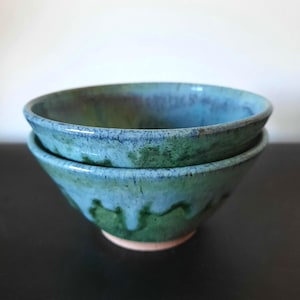 Può includere: Due ciotole in ceramica impilate. Hanno una glassa turchese e verde con un aspetto strutturato. I bordi sono blu scuro e la base rosa chiaro. Le ciotole sono su una superficie scura. Adatte per servire cibo.