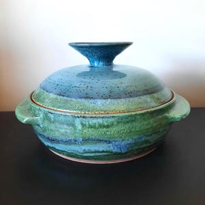 May include: A handmade ceramic casserole dish with a lid. The dish is glazed in shades of blue and green, with a darker blue handle on the lid. The dish has two side handles and a round shape.