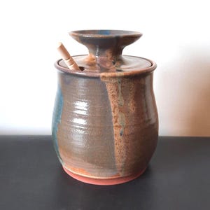 May include: A handcrafted ceramic jar with a lid and a small wooden spoon. The jar features brown, blue, and orange tones. The jar is placed on a black surface.