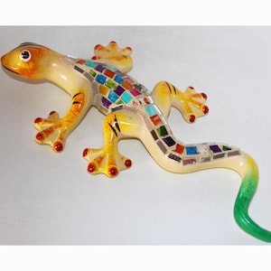 May include: A yellow ceramic gecko with a colorful mosaic pattern on its back and tail. The gecko has red eyes and red claws.