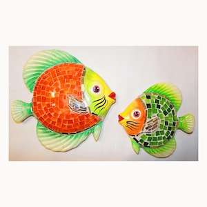 Puede incluir: Dos decoraciones de pared de peces de mosaico de colores. El pez más grande es naranja y verde con un ojo amarillo. El pez más pequeño es verde con un ojo amarillo y una boca naranja.