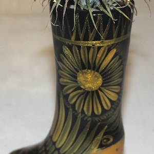 May include: A black and gold ceramic boot planter with a gold daisy design and an air plant growing inside.