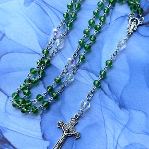 May include: A rosary with green beads and a silver crucifix. The beads are a vibrant green color, interspersed with clear, iridescent beads. The crucifix features a detailed depiction of Jesus Christ.