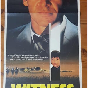 May include: Movie poster for the film "Witness" featuring Harrison Ford. The poster has a large image of Ford's face, with text at the top and bottom. The title "Witness" is in large yellow letters.