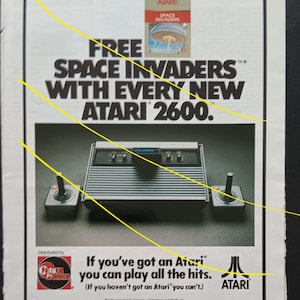 Op de afbeelding: Een advertentie voor de Atari 2600 videogameconsole. De advertentie toont de console, twee joysticks en de tekst "FREE SPACE INVADERS WITH EVERY NEW ATARI 2600." Het Atari-logo is ook zichtbaar.