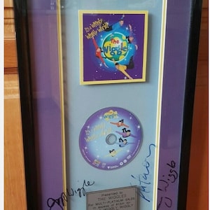 May include: Framed platinum sales award for The Wiggles' "It's a Wiggly Wiggly World" album. The frame includes a CD and album art, with signatures and a plaque. The frame is black, with purple and light blue accents.