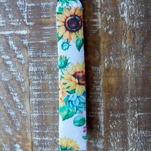 May include: A white key chain with a black strap and a silver key ring. The strap is covered in a sunflower print with yellow, green, and brown colors.