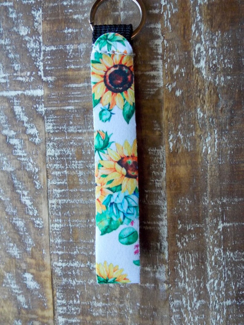 May include: A white key chain with a black strap and a silver key ring. The strap is covered in a sunflower print with yellow, green, and brown colors.