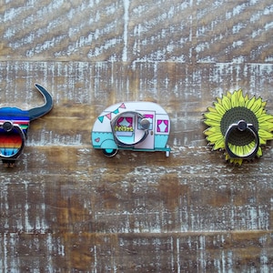 May include: Three phone ring holders with different designs. The first is a blue and white bull, the second is a light blue and pink camper, and the third is a yellow sunflower.